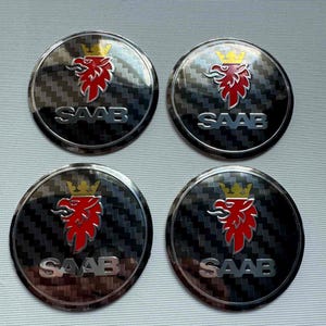 Könnte beinhalten: Vier runde Autoembleme mit einem Hintergrund aus Kohlefaser. Jedes Emblem zeigt einen roten Greif mit einer goldenen Krone und dem Wort "SAAB" in silbernen Buchstaben. Diese Embleme sind wahrscheinlich für ein Saab-Fahrzeug.