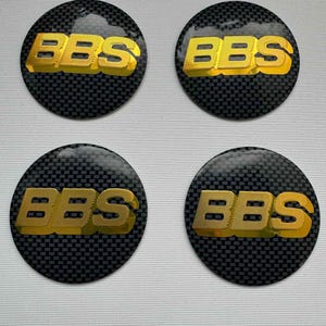 Może przedstawiać: Cztery okrągłe kołpaki kół samochodowych. Każdy kołpak ma złote litery "BBS" na czarnym tle z włókna węglowego. Kołpaki są ułożone w kwadrat na białym tle.