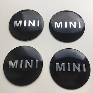 以下が含まれることがあります： 黒い円形の車のエンブレム4つ。銀色の文字で「MINI」と書かれています。エンブレムは光沢があり、耐久性のある素材で作られているようです。背景は明るいニュートラルカラーです。