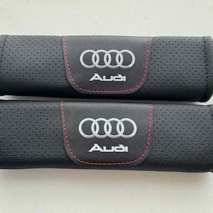 Könnte beinhalten: Zwei schwarze Sicherheitsgurtpolster mit dem Audi-Logo in Weiß und roten Nähten. Die Bezüge haben eine perforierte Textur, und das Audi-Logo ist auf einem schwarzen Patch zentriert.