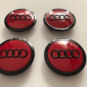 以下が含まれることがあります： 赤と黒の車のホイールセンターキャップ4個。各キャップには、アウディのロゴ、4つのインターロッキングリングのセットが、赤い背景に黒で表示されています。キャップは光沢仕上げで、黒い外側のリングが付いています。