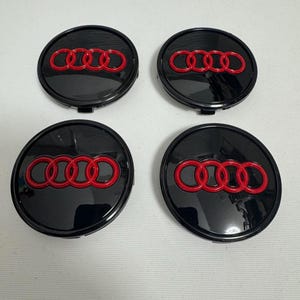 May include: Four black wheel center caps with a red Audi logo. The logo consists of four interlocking rings. The caps are round and have a glossy finish.