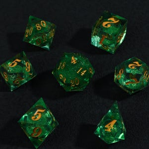 May include: Set of seven green and gold polyhedral dice for tabletop games. Each die is transparent with gold numbering and glitter, including a D4, D6, D8, D10, D12, D20, and a percentile die.