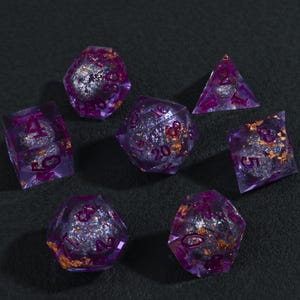 Puede incluir: Juego de siete dados poliédricos de varias formas. Los dados son de color púrpura translúcido con purpurina plateada y motas doradas. Los números están pintados en un púrpura más oscuro. Ideal para juegos de rol de mesa.