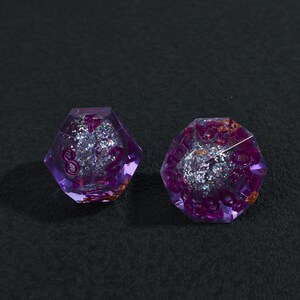 May include: Two purple, multi-sided dice with glitter and numbers visible inside. The dice are made of a clear material with purple edges and glittery silver and gold accents. The numbers are in a darker purple color.