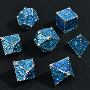 Op de afbeelding: Set van zeven blauwe en zilverkleurige polyhedrale dobbelstenen voor tabletop games. Elke dobbelsteen heeft een unieke vorm en nummer, met een metallic zilveren rand. De dobbelstenen hebben een getextureerde, glanzende afwerking en zijn gerangschikt op een donkere ondergrond.