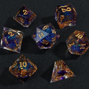Puede incluir: Un conjunto de siete dados poliédricos con un tema celestial. Los dados son transparentes con un interior de purpurina azul y morado y numeración dorada. Algunos dados tienen detalles de pan de oro.
