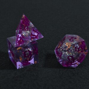 May include: Three purple resin dice with glitter and gold flakes. The dice are a cube, a pyramid, and a 20-sided die. The numbers are in a darker purple. The dice are for a tabletop game.