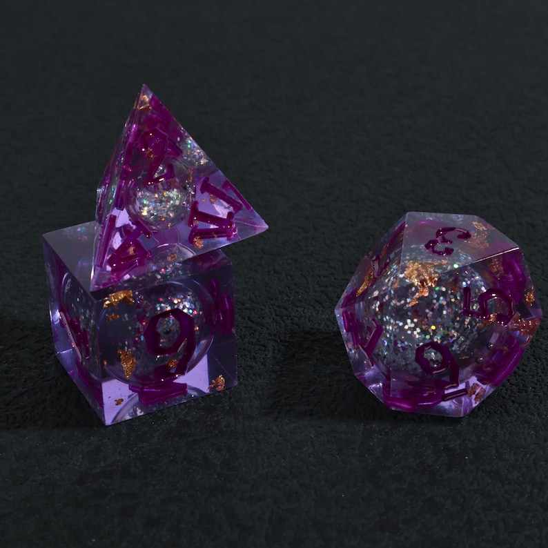 May include: Three purple resin dice with glitter and gold flakes. The dice are a cube, a pyramid, and a 20-sided die. The numbers are in a darker purple. The dice are for a tabletop game.
