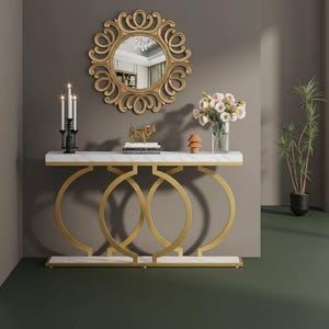 Op de afbeelding: Een goudkleurige console met een wit marmeren blad en een decoratieve cirkelvormige basis. Boven de tafel hangt een goudomrande spiegel, die is versierd met kaarsen, bloemen en decoratieve objecten. Een potplant staat op de achtergrond.