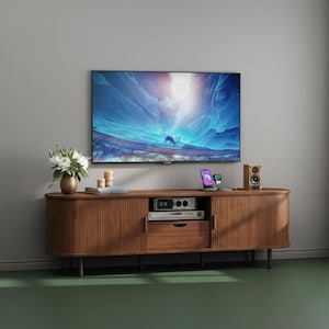 May include: A contemporary wooden TV cabinet with a large flat-screen television mounted above. The cabinet has a curved design with vertical grooves and black legs. A small speaker, a vase of flowers, and a smartphone are on the cabinet.