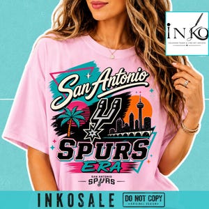 May include: A light pink t-shirt featuring a graphic print with the text San Antonio Spurs Era. The design includes a palm tree, a city skyline with a tower, a basketball-themed logo, and colorful geometric shapes in teal, orange, and black.