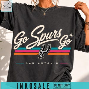 May include: A charcoal grey short-sleeve t-shirt featuring the text Go Spurs Go in a cream script font. Below the text are three horizontal stripes in teal, pink, and orange, a black Spurs logo, and the text SAN ANTONIO in a simple sans-serif font.