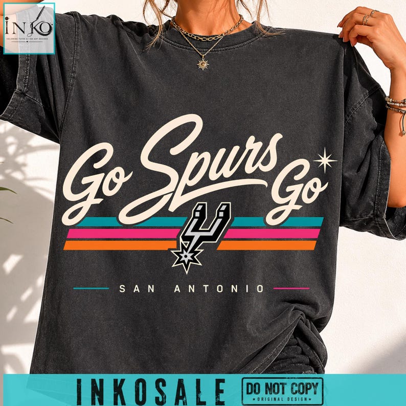 May include: A charcoal grey short-sleeve t-shirt featuring the text Go Spurs Go in a cream script font. Below the text are three horizontal stripes in teal, pink, and orange, a black Spurs logo, and the text SAN ANTONIO in a simple sans-serif font.