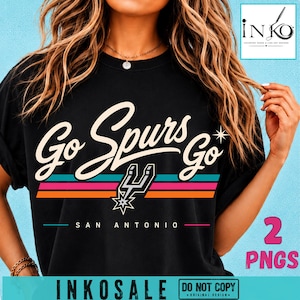 May include: A black t-shirt featuring the text Go Spurs Go in a cream script font, with a San Antonio Spurs logo and three horizontal stripes in teal, pink, and orange. The text SAN ANTONIO appears below the logo in a simple sans-serif font.