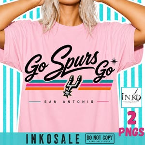 May include: A light pink t-shirt featuring the black text Go Spurs Go with a star icon and a San Antonio Spurs logo. Below the text are three horizontal stripes in teal, pink, and orange, with the words SAN ANTONIO printed in black below.
