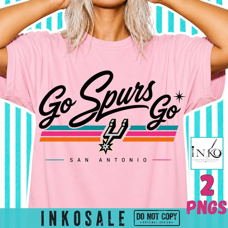 May include: A light pink t-shirt featuring the black text Go Spurs Go with a star icon and a San Antonio Spurs logo. Below the text are three horizontal stripes in teal, pink, and orange, with the words SAN ANTONIO printed in black below.