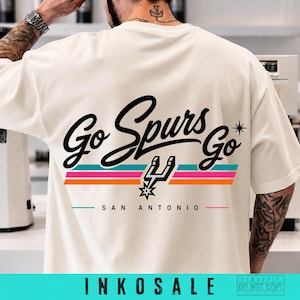 May include: An off-white short-sleeve t-shirt featuring the black cursive text Go Spurs Go on the back. Below the text are three horizontal stripes in teal, pink, and orange, a black San Antonio Spurs logo, and the text SAN ANTONIO in black.