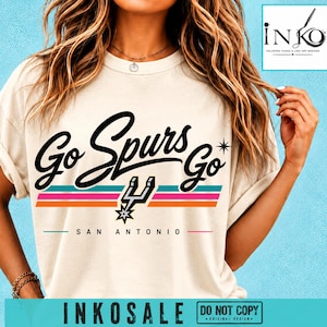 May include: A cream-colored t-shirt featuring the black text Go Spurs Go with a star icon, a San Antonio team logo, and horizontal stripes in teal, pink, and orange. The text San Antonio is printed below the graphic in a simple black font.