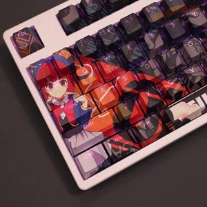 May include: A white mechanical keyboard with custom keycaps. The keycaps feature anime-style artwork with a red-haired character and abstract designs in red, orange, and black. The keyboard has standard letter and number keys.