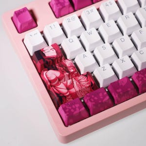 Peut inclure: Un clavier mécanique rose avec des touches blanches et magenta. Certaines touches présentent un motif de personnage d'anime rouge et rose. Le clavier a une disposition QWERTY standard.