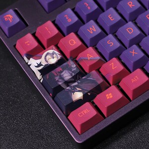 Può includere: Primo piano di una tastiera meccanica con tasti viola e magenta. Alcuni tasti presentano illustrazioni di personaggi in stile anime. I tasti sono disposti in un layout QWERTY standard, con lettere e simboli bianchi.