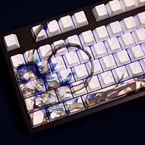 Puede incluir: Un teclado mecánico negro con teclas blancas. Algunas teclas presentan ilustraciones de estilo anime con detalles en azul y dorado. El teclado tiene una distribución estándar con teclas de función y un teclado numérico. Las teclas están retroiluminadas.
