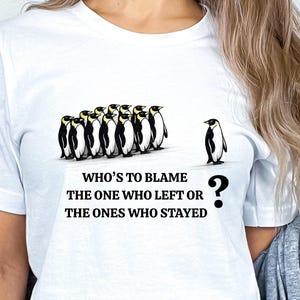 May include: White t-shirt featuring a graphic of penguins. A group of penguins stands together, while one penguin is isolated. The text reads, "WHO'S TO BLAME THE ONE WHO LEFT OR THE ONES WHO STAYED?" A question mark is next to the single penguin.