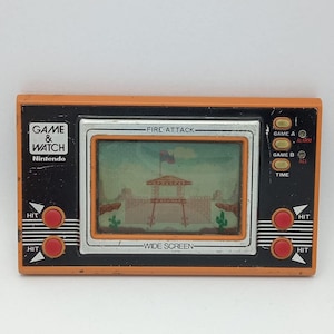 May include: An orange and black Nintendo Game & Watch handheld electronic game. The screen displays a western-themed game titled "Fire Attack." Features include red "HIT" buttons, game selection buttons, and a time function. The game is a "WIDE SCREEN" model.
