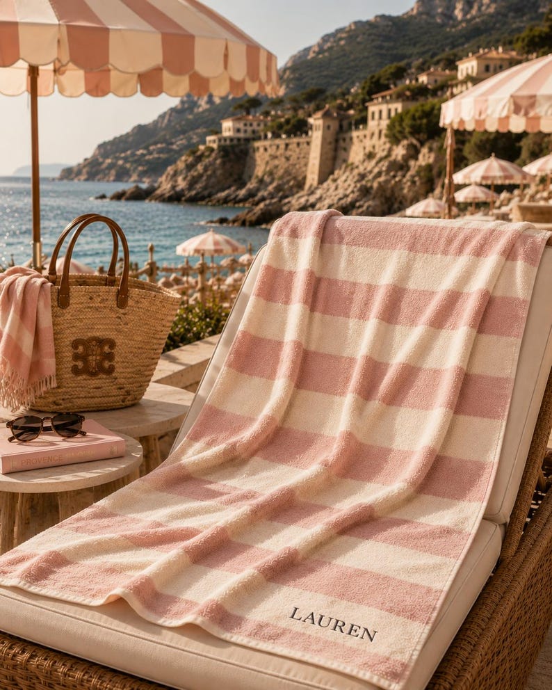 Op de afbeelding: Een strandsc&egrave;ne met een roze en wit gestreepte handdoek over een ligstoel. Een strooien tas en een boek liggen op een bijzettafel. Een gestreepte parasol biedt schaduw. Het woord "LAUREN" is op de handdoek geborduurd.