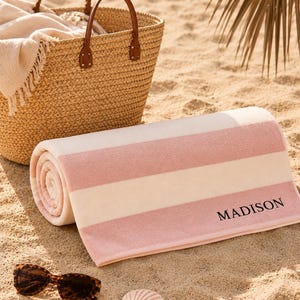 Op de afbeelding: Een opgerolde strandhanddoek met roze en witte strepen, gepersonaliseerd met de naam "MADISON" in zwarte letters. Een rieten mand met leren handvatten en een zonnebril liggen op het zand, wat duidt op een stranddag.