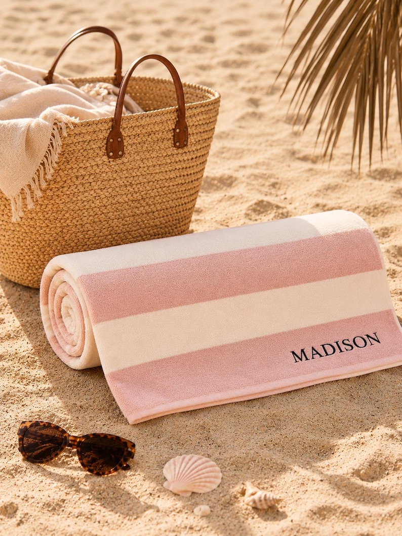 Op de afbeelding: Een opgerolde strandhanddoek met roze en witte strepen, gepersonaliseerd met de naam "MADISON" in zwarte letters. Een rieten mand met leren handvatten en een zonnebril liggen op het zand, wat duidt op een stranddag.
