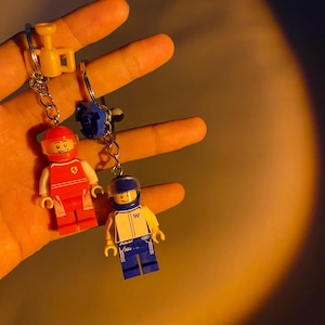 May include: Two Lego-style keychains featuring miniature race car driver figures. One figure is red with a white torso and a red helmet, the other is blue and yellow. The keychains are held by a silver chain and a yellow and blue accessory.