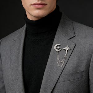 Puede incluir: Broche plateado con diseño de luna creciente y estrella, adornado con pequeños cristales brillantes. El broche está conectado por una delicada cadena. Se sujeta a una blazer gris, combinado con un cuello alto negro.