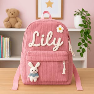 Puede incluir: Mochila de pana rosa con el nombre "Lily" en letras blancas cursivas. Cuenta con una flor blanca y un pequeño conejito con una bufanda azul. Incluye un bolsillo frontal con cremallera de borla blanca y correas rosas.