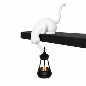 May include: A white cat figurine in a crouching position perched on the edge of a black shelf. The cat has its tail extended downward, holding a small black lantern with a glowing light inside. The design features a smooth matte finish.