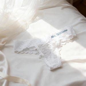 Peut inclure: String blanc en dentelle avec bord festonné et petit nœud. Le texte "Mrs. Williams" est brodé en fil bleu clair sur la ceinture. La lingerie est présentée sur une surface en satin blanc.