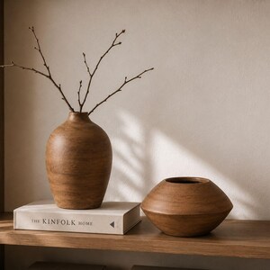 May include: Two brown wooden vases with a smooth finish. One vase is tall and rounded with thin bare branches inside, while the other is short and wide with a tapered shape. They sit on a white book titled The Kinfolk Home on a wooden shelf.