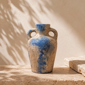 Könnte beinhalten: Eine Keramikvase mit einer texturierten, verwitterten beigen Oberfläche und Flecken aus leuchtend blauer Glasur. Das Gefäß hat einen abgerundeten Körper, einen schmalen Hals und zwei symmetrische Henkel, die von den Seiten zum Hals führen.