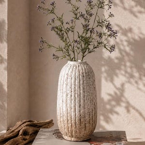 Könnte beinhalten: Eine hohe, zylindrische Keramikvase mit einer strukturierten, cremeweißen Oberfläche und vertikalen Rillen. Die Vase enthält ein Bündel dünner, grüner Zweige mit kleinen violetten Blüten. Sie steht auf einem rechteckigen Buch mit dem Titel VOGUE.