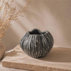 May include: A round, dark grey ceramic vase with a textured, ribbed surface featuring white distressed highlights. The vessel has a wide, open mouth and a bulbous, organic shape, showcasing a weathered finish across its exterior vertical ridges.