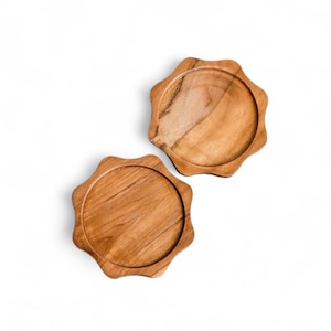 May include: Two wooden coasters with a unique, scalloped edge design. The coasters are made of a light brown wood, showcasing the natural grain. They are circular with a slightly raised edge, perfect for protecting surfaces from drink condensation.