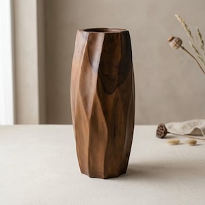 Puede incluir: Un jarrón de madera alto y cilíndrico con un acabado marrón oscuro y una superficie exterior geométrica y facetada. El jarrón tiene una abertura superior hueca y una estructura tallada sólida con patrones de veta de madera visibles en todo el cuerpo.