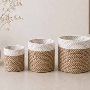 Puede incluir: Un conjunto de tres cestas de almacenamiento cilíndricas hechas de cuerda trenzada. Cada cesta presenta una sección inferior de color tostado natural y una sección superior blanca en contraste, dispuestas en orden de tamaño.