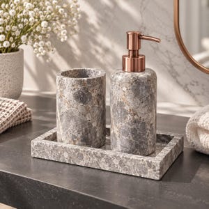 Puede incluir: Un juego de accesorios de baño que incluye un dispensador de jabón cilíndrico con bomba de color cobre, un vaso cilíndrico a juego y una bandeja rectangular. Las tres piezas están hechas de piedra gris con un patrón moteado y veteado.