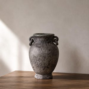 May include: A dark grey stone vase with a mottled, textured surface, a wide rounded body, a flared rim, and two small circular handles attached to the neck. The vessel features a pedestal base and a weathered, speckled appearance throughout.