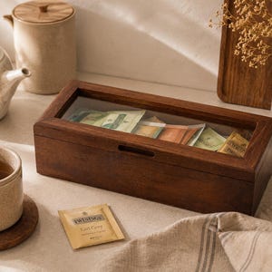 Könnte beinhalten: Eine rechteckige Tee-Aufbewahrungsbox aus dunkelbraunem Holz mit einem transparenten Glasdeckel, die mehrere Teebeutel enthält. Ein einzelner Twinings Earl Grey Teebeutel liegt vor der Box, die eine kleine horizontale Griffmulde besitzt.