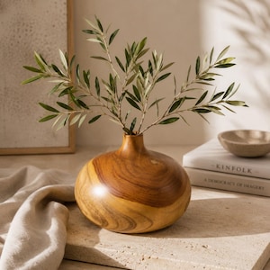 May include: A round, polished wooden vase with a wide base and a narrow neck, containing a sprig of green olive branches. The wood features prominent natural grain patterns in shades of light and dark brown, showcasing a smooth, rounded shape.