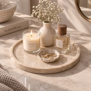 May include: A round beige marble tray holding a clear glass candle with a white label reading Santal, a small cream ceramic vase with white baby's breath flowers, a square perfume bottle with a wooden cap, and a small scalloped dish with gold jewellery.