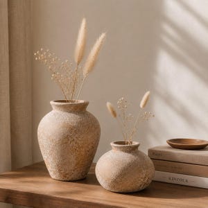 May include: Two round, textured beige ceramic vases with a rough, stone-like finish. The larger vase is taller and bulbous, while the smaller one is squat and rounded. Both contain dried beige grasses and delicate white baby's breath flowers.